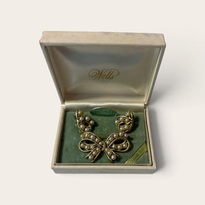 May include: A gold-toned metal brooch shaped like a bow, accented with small white pearls, presented in a cream-colored box with the Wells brand name printed on the interior lid and a green velvet insert holding the jewelry piece securely.