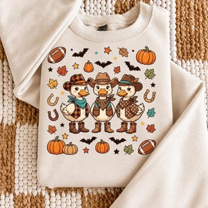 Pode incluir: Uma camisola bege de gola redonda com um gráfico de três patos a usar chapéus de cowboy, bandanas e botas. O design é rodeado por ícones de outono, incluindo abóboras, folhas de ácer, bolas de futebol americano, ferraduras e morcegos.