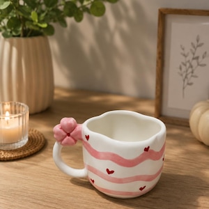 Può includere: Tazza in ceramica bianca con strisce rosa ondulate e piccoli cuori rossi. Il manico è a forma di fiore rosa. La tazza è su una superficie di legno, con una candela, una pianta e un'opera d'arte incorniciata sullo sfondo. Una tazza da caffè affascinante e decorativa.