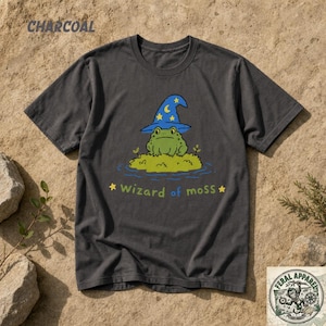 Puede incluir: Una camiseta gris carbón de manga corta con un gráfico de una rana verde con un sombrero de mago azul con estrellas amarillas. La rana está sentada sobre un parche de musgo en agua azul. Debajo, el texto dice Wizard of moss en amarillo.