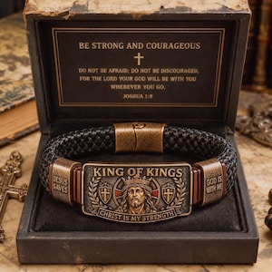 May include: A black braided leather bracelet with a rectangular bronze-toned metal centerpiece featuring an embossed image of Jesus, the text King of Kings, and Christ is My Strength, presented in a black box with the Joshua 1:9 scripture.