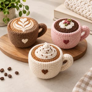 以下が含まれることがあります： コーヒーをテーマにしたデザインのクロッシェ編みのマグカップ3点。ラテアートが施された茶色のマグ、白い花とベリーがあしらわれたピンクのマグ、ホイップクリームがのったクリーム色のマグ。それぞれのマグの前面には小さなハートのディテールがあります。