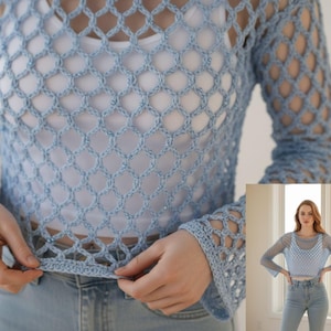 May include: A light blue long-sleeve crochet top featuring an open-knit diamond pattern with a solid hemline. The garment is worn over a plain white tank top and paired with light blue denim jeans, showcasing a textured, lightweight construction.