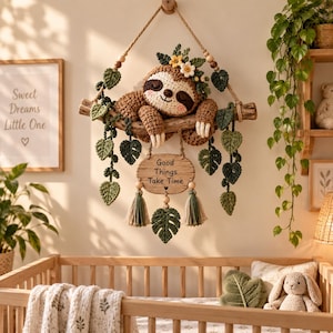 Può includere: Un bradipo all'uncinetto appeso a un ramo di legno, decorato con foglie di filato verde e piccoli fiori. Sotto il bradipo, un cartello in legno pende da una corda con il testo 'Good Things Take Time', due nappe di filato verde e una foglia.