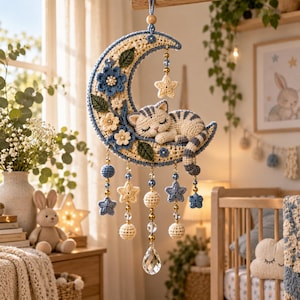 Peut inclure: Une suspension en crochet représentant un croissant de lune crème orné de fleurs bleues et de feuilles vertes. Un chat rayé gris et blanc dort sur la lune. Des étoiles bleues et crème, des perles et un cristal transparent pendent de la lune.