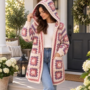 Könnte beinhalten: Eine langärmelige Kapuzenstrickjacke aus cremefarbenen, altrosa und lavendelfarbenen Häkel-Granny-Squares. Das Kleidungsstück hat eine offene Vorderseite, zwei große aufgesetzte Taschen und gerippte Bündchen und wird über einem weißen Oberteil getragen.
