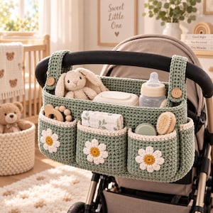 May include: A sage green crochet stroller organizer featuring three front pockets, each adorned with a white and yellow daisy applique. The organizer includes two wooden buttons for attachment and contains a plush bunny, a baby bottle, and a brush.
