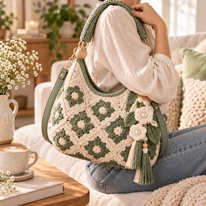 May include: A crochet hobo bag featuring a cream and sage green floral granny square pattern. The bag includes a sage green strap, gold-tone hardware, wooden bead accents, and a decorative charm with crochet flowers, leaves, and a long tassel.