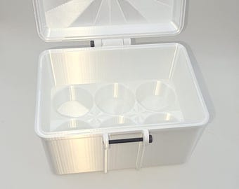 May include: A white rectangular storage box with an open hinged lid. The interior base features six circular indentations arranged in two rows of three. The lid has a sunburst pattern on the underside, and a black latch is visible on the front.