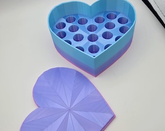 May include: A heart-shaped storage container with a removable lid. The base features a light blue exterior with a pink bottom edge and contains multiple circular slots for organizing items. The lid is light purple with a geometric starburst pattern.