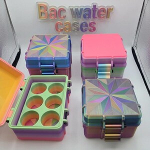 May include: Four square storage cases with the text Bac water cases displayed above. The cases feature vibrant, multi-colored gradient finishes, some with geometric starburst patterns on the lids. One case is open, revealing six circular interior compartments.