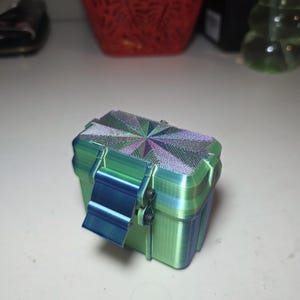 May include: A small, rectangular, 3D-printed storage container with a green and blue gradient finish. The lid features a textured, starburst-like geometric pattern in iridescent purple and green. A blue latch is secured with two black metal screws.
