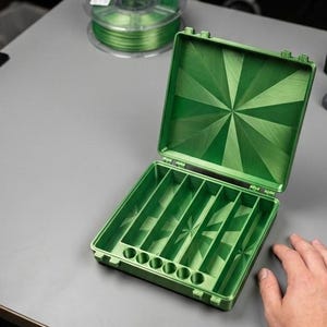 May include: A square, hinged storage case in a metallic green finish. The interior features five long, narrow rectangular compartments and a row of six small circular holes along the front edge. The lid interior displays a radiating sunburst pattern.