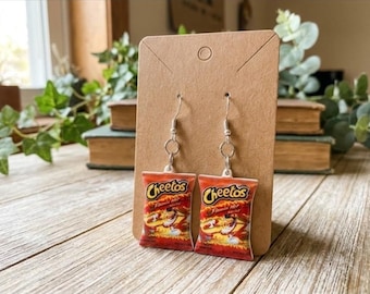 May include: A pair of dangle earrings featuring miniature replicas of Cheetos Flamin' Hot snack bags. The charms are red and orange with the Cheetos logo and text printed on the front, suspended from silver-toned metal earring hooks.