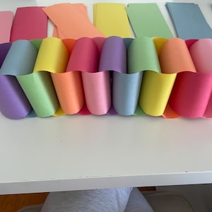 Puede incluir: Una cadena de papel multicolor construida con bucles entrelazados de cartulina en tonos morado, azul, verde, amarillo, naranja y rosa. La cadena está dispuesta en una fila horizontal, con tiras rectangulares sueltas de papel a juego.