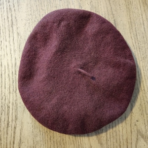 Könnte beinhalten: Eine runde, tiefburgunderrote Baskenmütze aus weichem, gefilztem Material. Die Kopfbedeckung hat eine glatte, kreisförmige Silhouette mit einem kleinen, mittig platzierten Stift an der Krone und weist eine gleichmäßige, dichte Textur auf.