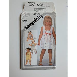 以下が含まれることがあります： 幼児用衣類のSimplicity 5910型紙封筒。表面には、ウエストに赤い紐が付いた白いノースリーブのドレスを着た子供の写真と、3種類の追加の衣装バリエーションのイラストが掲載されています。