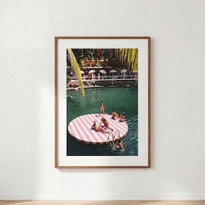 Op de afbeelding: Een ingelijste kunstdruk met een bovenaanzicht van een rond drijvend platform met roze en witte strepen op donkergroen water. Verschillende personen ontspannen zich op het platform, met palmbladeren en strandparasols op de achtergrond.