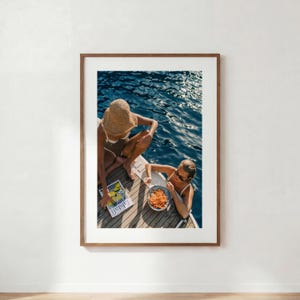 May include: A framed wall art print featuring two individuals on a wooden boat deck over dark blue water. One person wears a straw hat and swimsuit, holding a magazine titled Falstaff. The other person wears a white swimsuit and eats pasta.