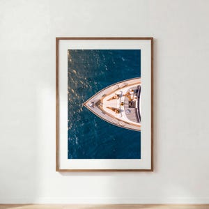 Könnte beinhalten: Ein gerahmter Wandkunstdruck mit einer Luftaufnahme des Bugs einer weißen Yacht auf tiefblauem Wasser. Das Yachtdeck ist mit gestreiften Kissen und zwei liegenden Figuren ausgestattet. Der Holzrahmen umgibt das vertikale rechteckige Foto.