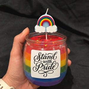 Pode incluir: Uma vela em frasco de vidro com camadas de cera nas cores do arco-íris e um rótulo branco com o texto preto Stand with Pride. O topo é preenchido com cera vermelha e glitter em forma de estrela, com um topo de vela de arco-íris e nuvens.