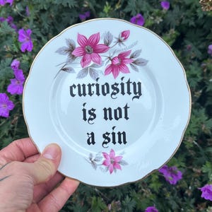 Pode incluir: Um prato de cerâmica branca com borda recortada dourada, apresentando ilustrações florais cor-de-rosa e o texto preto "curiosity is not a sin" impresso numa fonte de estilo gótico centrado na superfície do prato decorativo redondo.
