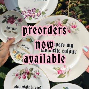 May include: A collection of five white ceramic plates with gold-trimmed edges, each featuring floral illustrations and black gothic-style text. The plates display phrases including 'i was your robot companion', 'right foot in the roses', 'left foot on a landmine', 'were my favourite colour', 'what might be good for your heart', and 'might not be good for my head'.