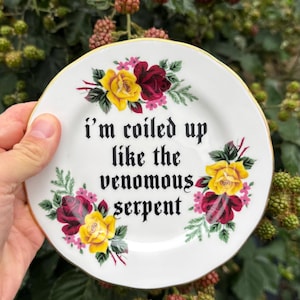 May include: A white ceramic plate with a gold-trimmed edge, featuring clusters of yellow and dark red roses with green leaves. The center displays the black text: i'm coiled up like the venomous serpent, written in a gothic-style typeface.