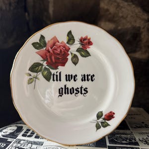 May include: A white ceramic plate with a scalloped gold rim, featuring a red rose illustration and the black text 'til we are ghosts' printed in a gothic font. The plate is decorated with a large rose cluster at the top and a single rose at the bottom.