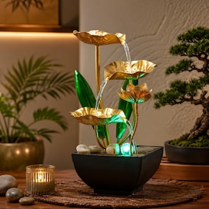 以下が含まれることがあります： 金色の蓮の葉と中央の花をあしらった卓上噴水。水は一番上の葉から下の段へと流れ落ち、黒い四角い水槽に溜まります。噴水には緑色の茎と、底面に光るライトが組み込まれています。