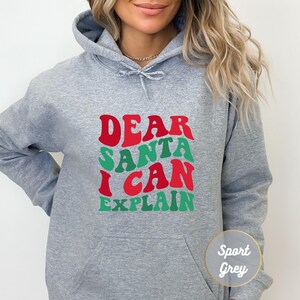 以下が含まれることがあります： レトロな波状のフォントで「DEAR SANTA I CAN EXPLAIN」という文字がプリントされたスポーツグレーのフード付きスウェットシャツ。DEARとCANの文字は赤色、SANTAとEXPLAINの文字は緑色です。前面にはカンガルーポケットが付いています。