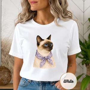 Puede incluir: Una camiseta blanca de manga corta con una ilustración gráfica de un gato siamés de ojos azul brillante. El gato lleva un pañuelo de color violeta claro con un pequeño estampado floral blanco alrededor del cuello. La camiseta tiene cuello redondo.