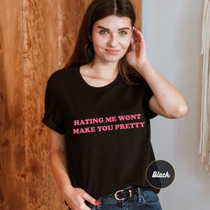 Op de afbeelding: Een zwart T-shirt met korte mouwen en de tekst "HATING ME WONT MAKE YOU PRETTY" in roze letters op de borst. Het shirt heeft een ronde hals en een standaard pasvorm, met een klein rond label dat de kleur zwart aangeeft.