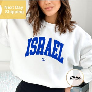 May include: A white long-sleeve crewneck sweatshirt featuring the word ISRAEL printed in a bold, arched blue font across the chest, with a small blue and white Star of David flag icon centered directly below the text.