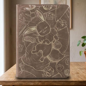 Op de afbeelding: Een rechthoekige bruine tablethoes met een in laser gegraveerde illustratie van een slapende Eevee, omringd door diverse Pokémon-elementen, waaronder een Poké Ball, een kleine fotolijst en gestileerde bloemmotieven op het oppervlak.