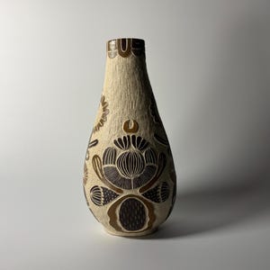 Puede incluir: Un jarrón de cerámica con una superficie texturizada de color crema que presenta patrones florales y geométricos tallados en marrón oscuro. El recipiente tiene un cuello estrecho y un cuerpo redondeado con detalles grabados.