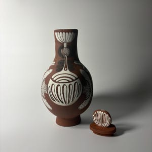 Puede incluir: Una vasija de cerámica color terracota con cuerpo redondeado y cuello estrecho, decorada con patrones botánicos pintados a mano en blanco y negro. Una tapa circular a juego con un diseño lineal en blanco y negro se sitúa al lado.