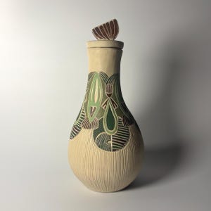 Puede incluir: Un recipiente de cerámica alto en forma de pera con una tapa a juego que presenta un asa en forma de abanico. El cuerpo es de color crema con una base texturizada de líneas verticales y una sección superior con patrones de gotas.