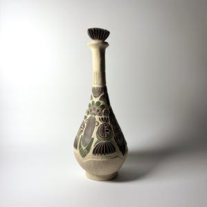 Puede incluir: Una jarra de cerámica beige, alta y esbelta, con cuello estrecho y tapón en forma de abanico. El cuerpo presenta patrones tallados en bajorrelieve de color marrón oscuro y verde apagado, con formas geométricas y motivos florales.