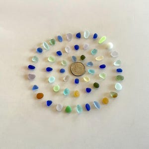 May include: A collection of small, smooth, multi-colored sea glass pieces arranged in a spiral pattern around a single silver-colored United States dime coin placed in the center, featuring shades of blue, green, white, yellow, and brown.