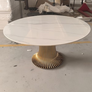 May include: A round dining table featuring a white tabletop with subtle grey veining patterns. The table is supported by a central pedestal base constructed from numerous vertical, curved gold-toned metal rods that flare out at the bottom.