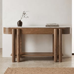 May include: A console table featuring a white marble top with grey veining and a medium-brown wooden base. The structure includes a central drawer, a lower shelf, and four cylindrical legs. A dark brown vase with a branch and two books sit on top.