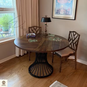 May include: A round wooden dining table with a dark brown finish and a black metal pedestal base featuring vertical slats. Two bamboo-style chairs with beige upholstered seats are positioned at the table, which holds a puzzle box and pieces.