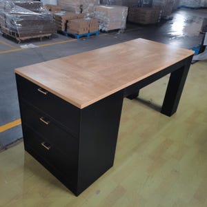 May include: A rectangular desk featuring a light brown wood grain top and a black base. The left side of the desk includes three stacked drawers, each equipped with a thin, horizontal metallic handle. The desk has a sturdy, clean-lined design.