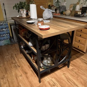 May include: A kitchen island featuring a rectangular wood-grain top and a black metal frame with circular side accents. The unit includes two open storage shelves below the main surface, providing space for various kitchen items and containers.
