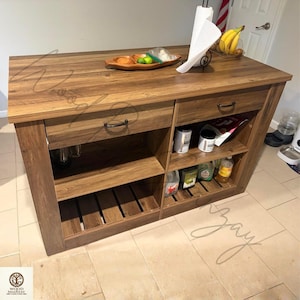 May include: A rectangular kitchen island with a dark brown wood grain finish, featuring two top drawers with black metal handles, two open storage shelves, and a slatted bottom shelf. The island has a flat countertop surface.