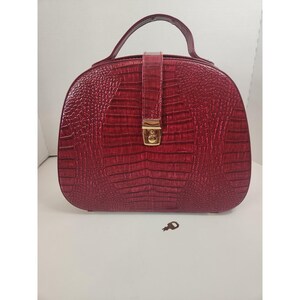 May include: A deep red handbag with a textured crocodile skin pattern, featuring a single top handle, a vertical central strap, and a gold-toned metal clasp closure. A small, dark, key-shaped object is positioned separately below the handbag.