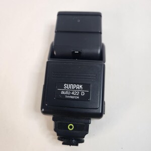 May include: A black Sunpak auto 422 D Thyristor camera flash unit. The device features a rectangular head, a central control body with the brand name and model printed in white text, and a mounting base with a small green circular indicator.