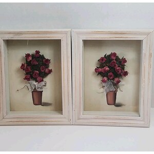 Può includere: Una coppia di cornici a cassetta con bordi in legno bianco anticato. Ogni cornice contiene un'opera d'arte tridimensionale che raffigura un vaso color terracotta con un mazzo di rose rosa scuro e un nastro bianco trasparente alla base.