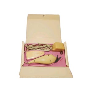 May include: A vintage electric fabric cutter with a cream-colored plastic body and a power cord, stored in a light beige folding case with a pink interior tray. The set includes a separate cream-colored power adapter and a protective plastic cover.
